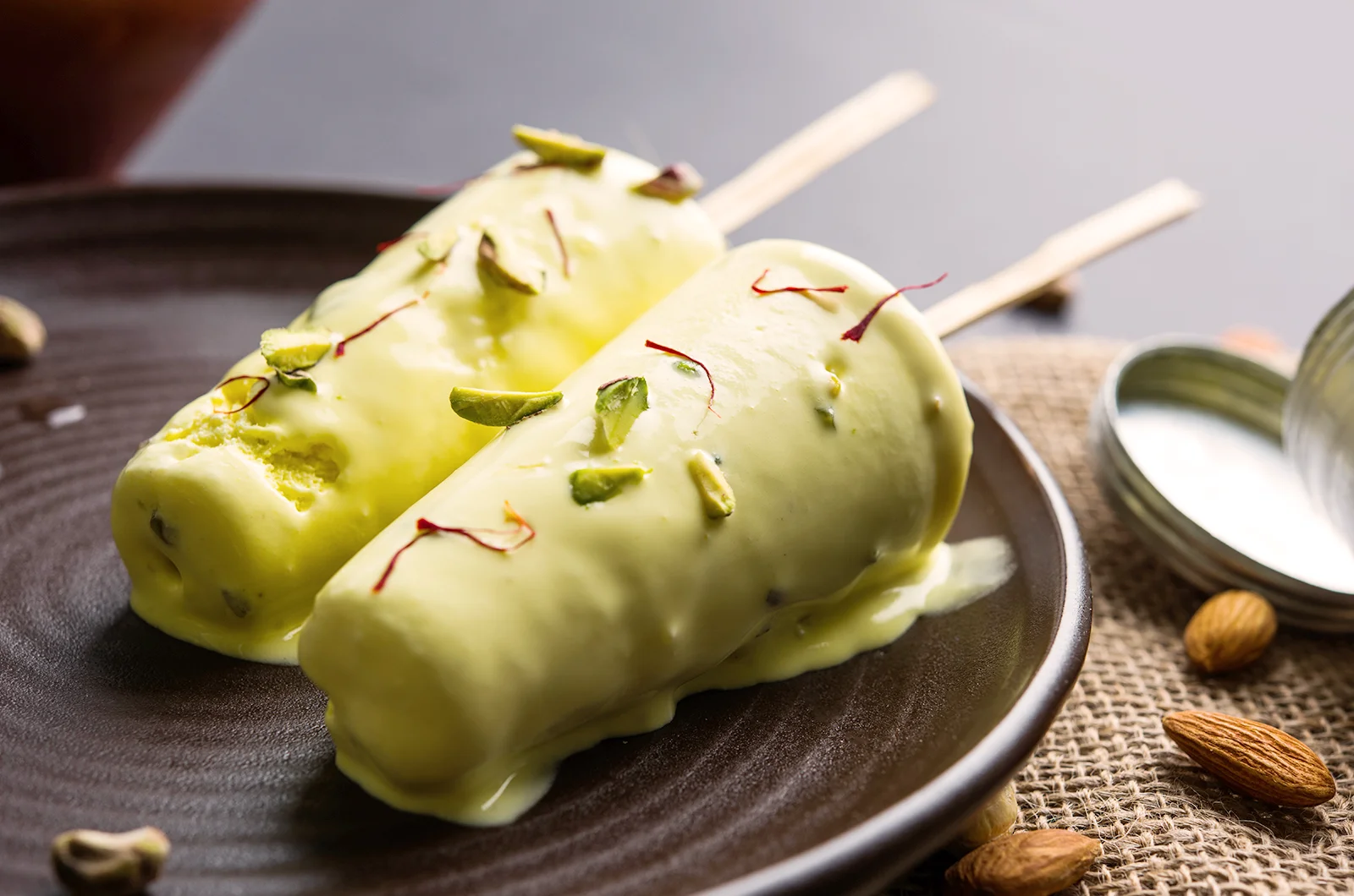 Two pistachio kulfi ice cream bars garnished with nuts and saffron on a dark plate.
